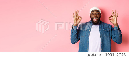 Impressed Black man in beanie and shirt, showing ok signs and staring at camera amazed, smiling happy, praise great offer, recommending product, pink background 103142798