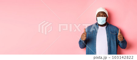 Covid-19, lifestyle and quarantine concept. Satisfied Black man in face mask showing thumbs-up, standing over pink background 103142822
