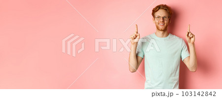 Attractive young man in glasses and t-shirt showing promo deal, pointing fingers up and smiling at camera, standing over pink background 103142842