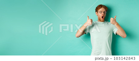 Excited young man with red hair and beard, staring at promo with awe, showing thumbs-up in approval, praising offer, standing over turquoise background 103142843