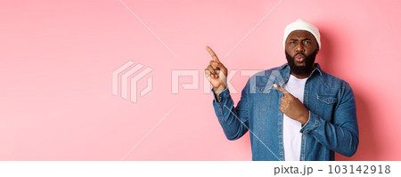 Confused and displeased african american man grimacing, cringe from disappointment and pointing fingers at upper left corner, bad promo, pink background 103142918