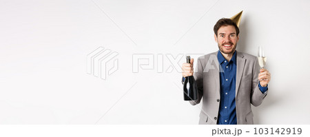 Celebration and holidays concept. Excited handsome birthday guy in party hat smiling, drinking champagne and having fun, white background 103142919
