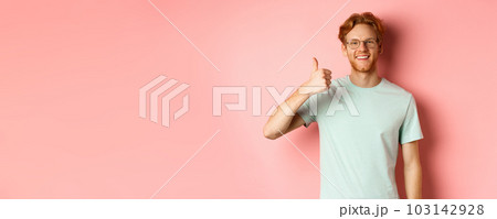 Cheerful redhead man in glasses and t-shirt, showing thumbs up with satisfied face, showing positive reaction, approve and agree with you, standing over pink background 103142928