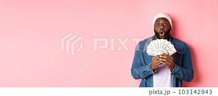 Amazed african-american man receive cash prize, showing money and staring at camera in awe, standing over pink background 103142943