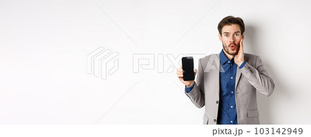 E-commerce and online shopping concept. Excited man gasping amaze and showing empty smartphone screen, wearing business suit, white background E-commerce and online shopping concept. Excited man gasping amaze and showing empty smartphone screen, wearing business suit, white background 103142949