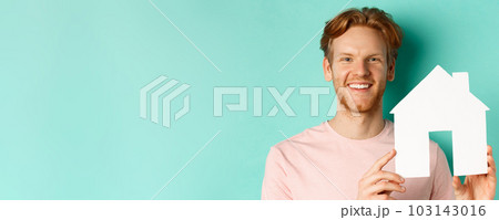 Real estate concept. Close up of redhead bearded man in t-shirt showing paper house cutout, smiling happy at camera, standing over turquoise background Real estate concept. Close up of redhead bearded man in t-shirt showing paper house cutout, smiling happy at camera, standing over turquoise background 103143016