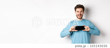 E-commerce and shopping concept. Smiling cheerful man showing empty smartphone screen in horizontal position, looking happy at camera, white background E-commerce and shopping concept. Smiling cheerful man showing empty smartphone screen in horizontal position, looking happy at camera, white background 103143030