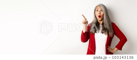 Impressed korean woman with red lips and trendy blazer, pointing and looking upper left corner, showing awesome promo offer, standing over white background Impressed korean woman with red lips and trendy blazer, pointing and looking upper left corner, showing awesome promo offer, standing over white background 103143136