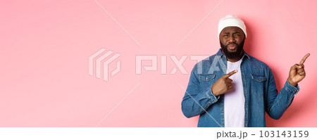Disappointed and skeptical african-american guy complaining, cringe from disgust, frowning and grimacing from dissatisfaction, pointing fingers right, pink background 103143159