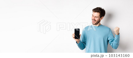 E-commerce and shopping concept. Lucky man winning online, showing empty smartphone screen and triumphing, saying yes with fist pump, standing over white background 103143160