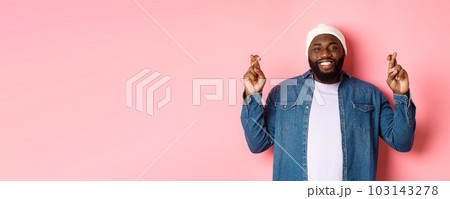 Optimistic african-american man making wish, holding fingers crossed and smiling, standing over pink background 103143278