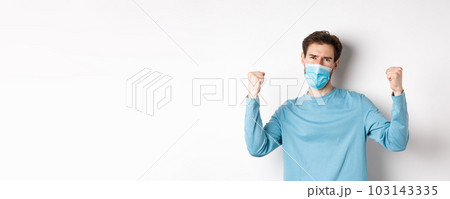 Covid-19, pandemic and social distancing concept. Happy young man in medical mask winning, screaming yes with satisfaction and raising hands up, celebrating victory, white background 103143335