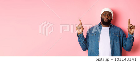Upset and gloomy african american man in beanie, pointing fingers up, sighing and looking at top with sadness, standing over pink background 103143341