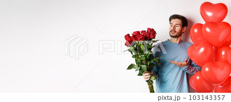 Romantic guy showing bouquet of red roses, pointing at flowers and standing near hearts balloons on Valentines day, prepare gifts for his lover, white background 103143357