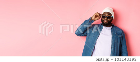 Cool and sassy african-american bearded man, looking confident, touching sunglasses and stare at camera please, standing over pink background 103143395