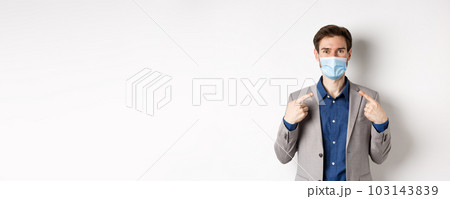 Covid-19, pandemic and business concept. Excited businessman in suit pointing at his medical mask and smiling with eyes, white background 103143839