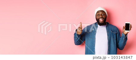 Online shopping and technology concept. Happy african-american hipster man showing thumbs-up and mobile screen, praising app, standing over pink background Online shopping and technology concept. Happy african-american hipster man showing thumbs-up and mobile screen, praising app, standing over pink background 103143847
