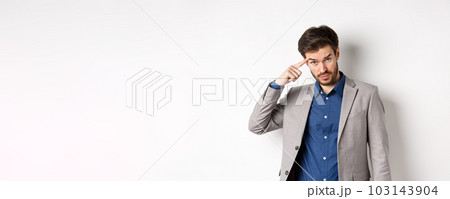 Annoyed male employer scolding personal, pointing finger at head, are you stupid out of your mind gesture, stare at camera from under forehead, standing in suit on white background 103143904