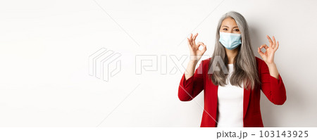 Coronavirus and business concept. Asian female manager in face mask looking cheerful, showing okay signs in approval, white background Coronavirus and business concept. Asian female manager in face mask looking cheerful, showing okay signs in approval, white background 103143925