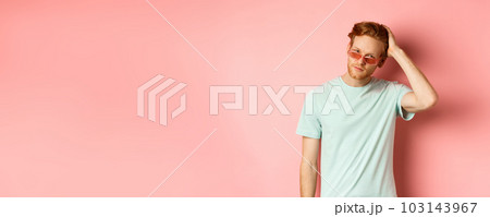 Handsome young redhead man in sunglasses, brushing hair with hand and looking smug and confident at camera, standing over pink background 103143967