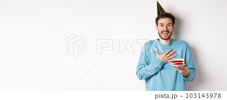 Celebration and holidays concept. Surprised birthday boy in party hat, holding bday cake and looking thankful, standing over white background Celebration and holidays concept. Surprised birthday boy in party hat, holding bday cake and looking thankful, standing over white background 103143978