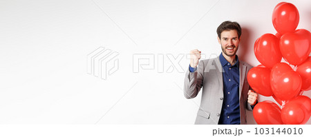 Lovers day. Cheerful young man in suit, feeling confident about Valentines date, saying yes and smiling, standing near hearts balloon, white background 103144010