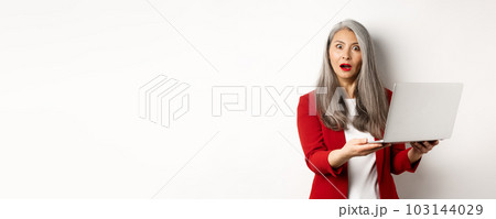 Business. Impressed asian old woman working on laptop, staring at camera surprised, standing over white background 103144029