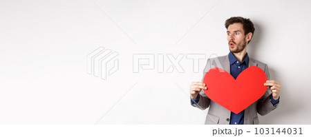 Man looking amazed at someone beautiful, holding big red heart cutout, standing on Valentines day over white background Man looking amazed at someone beautiful, holding big red heart cutout, standing on Valentines day over white background 103144031