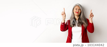 Skeptical and disappointed asian businesswoman pointing fingers up, grimacing and showing bad promo offer, standing over white background 103144042