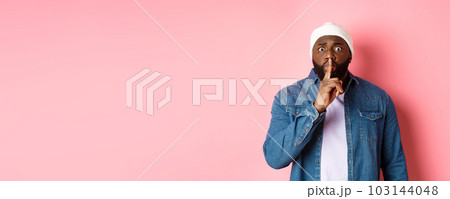 Worried Black man asking to keep quiet, sharing secret and hushing at you, holding finger pressed to lips and staring nervously at camera, pink background Worried Black man asking to keep quiet, sharing secret and hushing at you, holding finger pressed to lips and staring nervously at camera, pink background 103144048