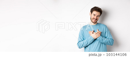 Portrait of young handsome man with beard, holding hands on heart and saying thank you, thinking about pleasant moment, standing over white background 103144106