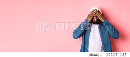 Happy Black man waiting for surprise, cover eyes and smiling, playing peekaboo, standing over pink background Happy Black man waiting for surprise, cover eyes and smiling, playing peekaboo, standing over pink background 103144135