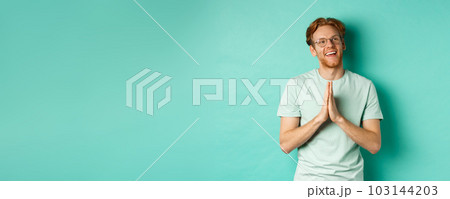 Hopeful redhead man with beard, wearing glasses and t-shirt, holding hands in namaste or plead gesture and looking right, smiling and thanking, standing over turquoise background 103144203