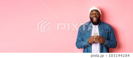 Technology and online shopping concept. Surprised young Black man using mobile phone, looking at camera amazed and happy after reading message, pink background 103144284