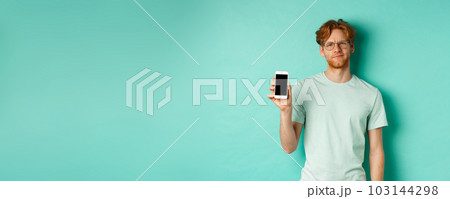 Skeptical male model with red hair and glasses, showing mobile screen and frowning disappointed, dislike application, standing over turquoise background 103144298