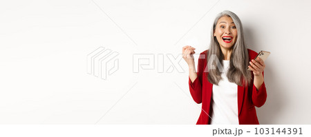 Excited asian businesswoman in red blazer, holding plastic credit card and smartphone, smiling happy at camera, standing over white background Excited asian businesswoman in red blazer, holding plastic credit card and smartphone, smiling happy at camera, standing over white background 103144391