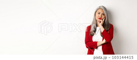 Annoyed and bothered asian businesswoman in red blazer, pouting and looking irritated at camera, standing over white background Annoyed and bothered asian businesswoman in red blazer, pouting and looking irritated at camera, standing over white background 103144415