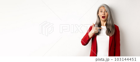 Surprised asian woman with grey hair, pointing at herself and gasping confused, standing over white background Surprised asian woman with grey hair, pointing at herself and gasping confused, standing over white background 103144451