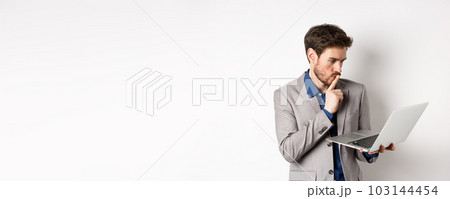 Pensive businessman working on laptop and thinking, looking thoughtful at computer screen, standing in suit on white background Pensive businessman working on laptop and thinking, looking thoughtful at computer screen, standing in suit on white background 103144454
