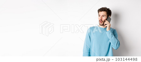 Technology concept. Intrigued young man looking left impressed, talking on mobile phone, calling someone, standing over white background Technology concept. Intrigued young man looking left impressed, talking on mobile phone, calling someone, standing over white background 103144498