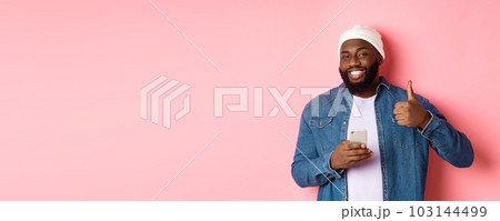 Technology and online shopping concept. Happy african-american guy showing thumb-up in approval while using mobile phone, standing satisfied over pink background 103144499