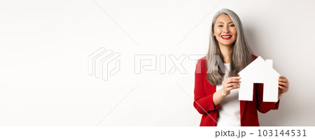 Asian female real estate agent showing paper house cutout, broker smiling friendly and selling property, standing over white background Asian female real estate agent showing paper house cutout, broker smiling friendly and selling property, standing over white background 103144531