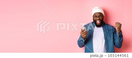 Technology and online shopping concept. Happy Black man rejoicing, winning in app, holding smartphone and scream yes, standing over pink background Technology and online shopping concept. Happy Black man rejoicing, winning in app, holding smartphone and scream yes, standing over pink background 103144561