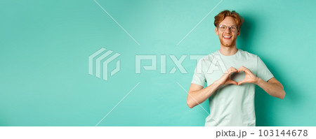 Valentines day and relationship concept. Happy boyfriend with red hair and beard, wearing glasses and t-shirt, showing heart sign and saying I love you, standing over turquoise background Valentines day and relationship concept. Happy boyfriend with red hair and beard, wearing glasses and t-shirt, showing heart sign and saying I love you, standing over turquoise background 103144678