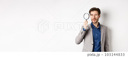 Handsome male model in suit looking through magnifying glass with interest, seeing something aside, white background Handsome male model in suit looking through magnifying glass with interest, seeing something aside, white background 103144833