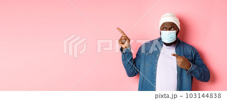 Coronavirus, lifestyle and global pandemic concept. Confused and disappointed african-american guy in face mask pointing fingers left, staring upset with camera, pink background 103144838