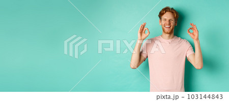 Attractive young man in t-shirt smiling satisfied, nod in approval and showing OK sign, approve and agree with something cool, standing over turquoise background Attractive young man in t-shirt smiling satisfied, nod in approval and showing OK sign, approve and agree with something cool, standing over turquoise background 103144843