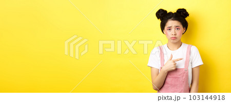 Sad and disappointed asian girl frowning, feel unfair, pointing finger right at bad thing, complaining on something upsetting, yellow background Sad and disappointed asian girl frowning, feel unfair, pointing finger right at bad thing, complaining on something upsetting, yellow background 103144918