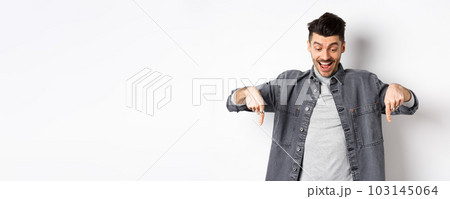 Excited european man looking and pointing fingers down, looking happy at advertisement, standing in denim jacket on white background 103145064