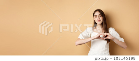 Valentines day and romance concept. Romantic caucasian girl in white dress showing heart gesture and smiling, say I love you, standing over beige background 103145199